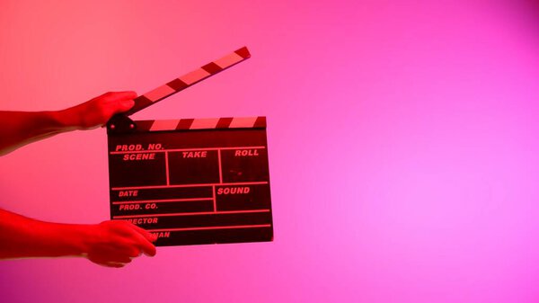 Hand is holding Black clapper board or movie slate on a pink background with a circular light, illuminated with red neon light. Cinema industry, entertainment