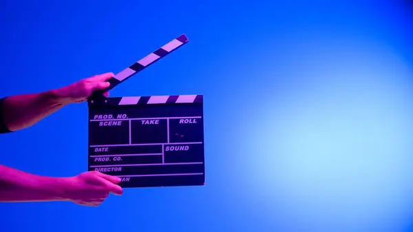 Hand is holding Black clapper board or movie slate on a blue background with a circular light, illuminated with pink neon light. Cinema industry, entertainment