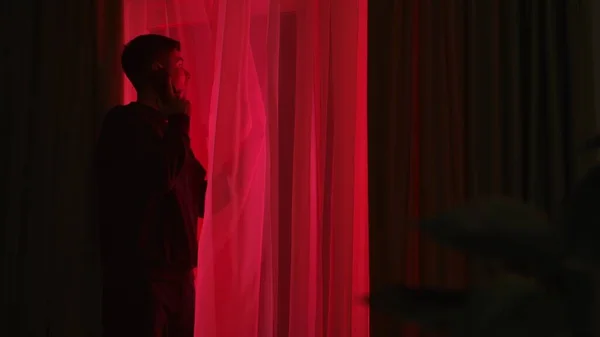 スマートフォンで話し 赤い警察のライトを窓から眺める男 窓の外を眺めている暗いアパートの奇妙な男 — ストック写真