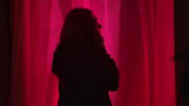 Kırmızı ve mavi polis ışıklarıyla aydınlatılmış karanlık bir odada pencerenin yanında duran bir kadının arka görüntüsü. Yaklaş. Endişeli kadın akıllı telefonuyla konuşuyor, endişeyle kafasını tutuyor ve...
