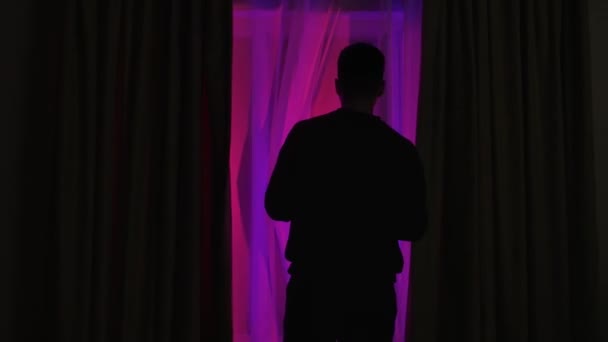 カーテンを暗い部屋で開け 窓の外で何が起こっているかを見る男の背中を見ます 点滅する警察のライトを見ている男 — ストック動画