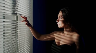 Yaratıcı vücut tanıtım konseptinin güzelliği. Stüdyoda güzel bir kadın modelin portresi. Camın yanında duran çekici kadın silueti jaluzinin yansımasıyla parmaklarıyla açar..
