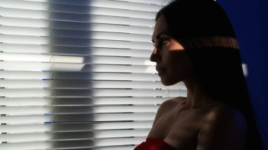 Yaratıcı vücut tanıtım konseptinin güzelliği. Stüdyoda kadın modelin portresi var. Kırmızı elbiseli çekici bir kadın pencerenin yanında dikilip, gözleri parlayan bir külüstürle dışarı bakıyor..