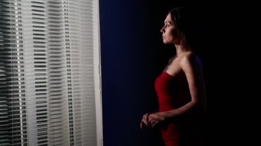 Vücut estetiği yaratıcı reklam konseptinin güzelliği. Stüdyoda çekici bir kadın modelin portresi. Kırmızı elbiseli esmer kadın pencerenin yanında dikilip mavi ışıktaki kameraya bakıyor..