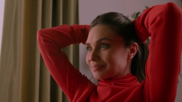 人类极端的情感创意广告概念 有魅力的女性模特儿在积极向上的形象 穿着红色高领毛衣的布鲁内特女人坐在房间里 笑着挽着头发 — 图库视频影像