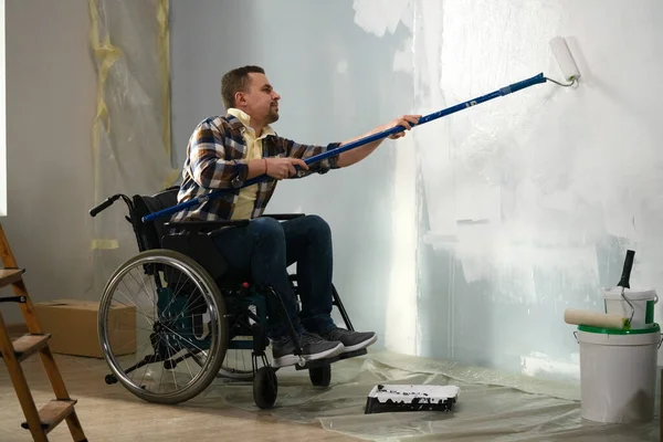The photo shows the room in which the repair is carried out. An adult man in a wheelchair. He is a repairman who draws a roller on a long stick of paint on the wall.