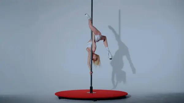 A young woman in white top and shorts dances pole dance choreography. Dancer in high-heeled boots erotically dancing in studio on white background