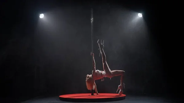 Modern dance style and choreography creative concept. Young female dancer in the studio. Professional pole dancer girl spinning on pylon, showing modern pole performance, black background with smoke.