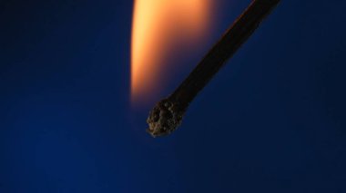 Mavi bir stüdyonun arka planında yanan bir kibritin makro çekimi. Yanan kibritin alevi karanlık uzayı aydınlatıyor. Yanan kibrit turuncu bir alevle kaplıdır.