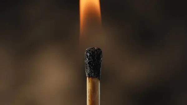 Macro shot of a burning match against a dark studio background. The flame of the burning match illuminates the dark space. The burning match is enveloped in an orange flame