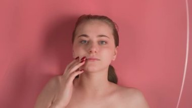 Pembe arka plan makyajsız genç ve çekici bir kızı gösteriyor. Görünüşte suyun altında yatıyor manikürlü bir elle yüzünü okşuyor ve kameraya bakıyor. Su dalgalar yaratırken.