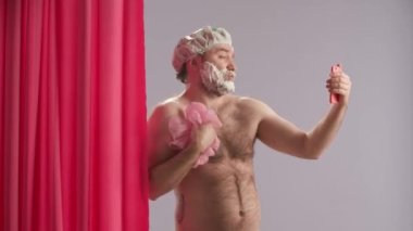 Su geçirmez başlık takan ve yüzünde tıraş köpüğü olan çıplak bir adam duş alıyor ve akıllı telefon kullanıyor. Adam selfie çekiyor, video çekiyor, canlı yayın yapıyor, pembe duş perdesinin yanında blog yazıyor.