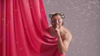 Seminer adamı, pembe duş perdesiyle kaplı, mavi arka planda, sabun köpükleriyle çevrili. Kafasında bigudileri ve gözlerinin altında hidrojel yamaları olan bir adam parmağını ağzına dayar.