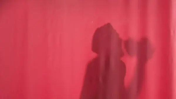 Silhouette of a man washing behind a pink curtain. Man in waterproof cap washing, dancing and singing, using loofah as a microphone. Body washing