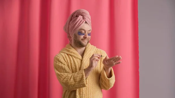 A man is grooming his nails after a shower, applying nail polish, nail oil with a brush. Man in yellow robe, with towel on his head and with hydrogel patches under his eyes in studio against pink
