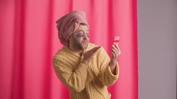 黄色いローブの男性 頭にピンクのタオル 目の下にヒドロゲルパッチが付いている男性は シャワーの後にスマートフォンを使用します 男はビデオ通話で話し 空気キスを送信し さよならを波する — ストック動画