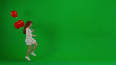 Çocukların yaratıcı konsepti. Stüdyoda küçük bir kızın portresi. Yeşil ekranda kırmızı helyum balonları olan beyaz elbiseli küçük bir kız. Kız kadraja giriyor.