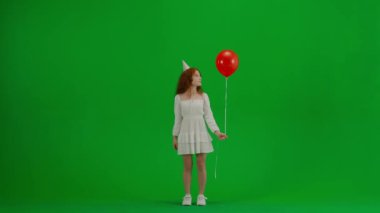 Çocukların yaratıcı konsepti. Stüdyoda küçük bir kızın portresi. Kırmızı helyum balonlu beyaz elbiseli küçük bir kız. Krom anahtar yeşil ekran. Parti konisi şapkasında izole edilmiş bir arka plan..