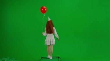 Beyaz elbiseli, kafasında tatil şapkası olan, yeşil arka planda kırmızı balonlarla yürüyen kızıl saçlı küçük bir kız. Arkadan bak. Tatil, neşe ve eğlence kavramı