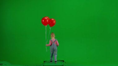 Çocukların yaratıcı konsepti. Stüdyoda küçük bir kızın portresi. Kot pantolonlu küçük bir kız ve kırmızı helyum balonlu bir at kuyruğu. Krom anahtar yeşil ekranda yürüyor. Arka plan izole edilmiş..