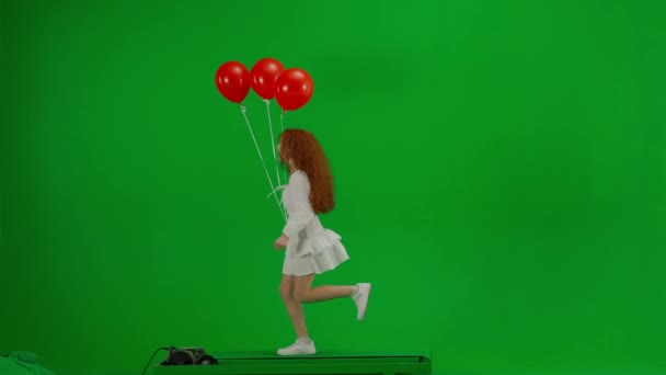スタジオの緑の背景に赤い風船で走る白いドレスで赤い女の子を斬首しました サイドビュー 休日のコンセプト 喜びと楽しみ — ストック動画