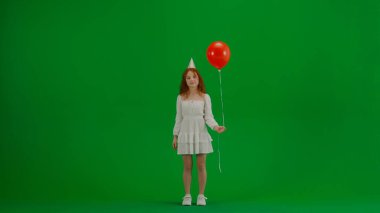 Çocukların yaratıcı konsepti. Stüdyoda küçük bir kızın portresi. Kırmızı helyum balonlu beyaz elbiseli küçük bir kız. Krom anahtar yeşil ekran. Parti konisi şapkasında izole edilmiş bir arka plan..