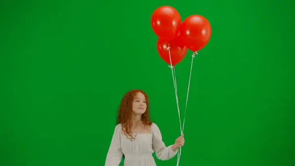 儿童创意概念 工作室里小女孩的画像 身穿白色长裙的小女孩 戴着红色气球 在彩色键绿色屏风上独立行走 看着相机 正面朝前看 — 图库照片