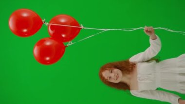 Stüdyoda küçük bir kızın portresi. Krom anahtar yeşil ekranında kırmızı helyum balonları olan beyaz elbiseli küçük bir kız parmak uçlarında durmuş gülümseyen yüze bakıyor. Dikey video.