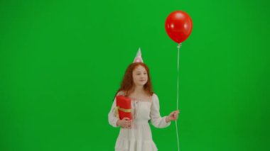 Çocukların yaratıcı konsepti. Stüdyoda küçük bir kızın portresi. Kırmızı balon renkli, yeşil ekranlı, beyaz elbiseli küçük kız parti şapkalı, hediye kutusuyla etrafta dolaşıyor..