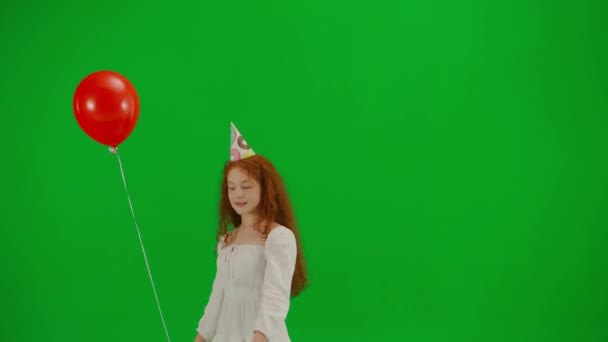 身穿白衣头戴节日帽的红头发小女孩头戴红色气球 站在工作室的绿色背景上 欢乐和欢乐的概念 — 图库视频影像