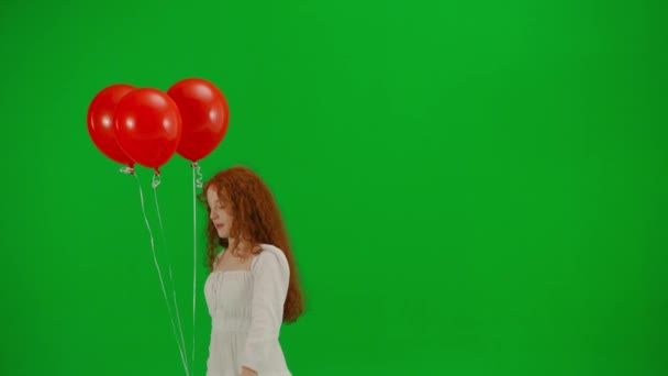 スタジオの緑の背景に赤い風船で走る白いドレスで赤い女の子を斬首しました ハーフターン 休日のコンセプト 喜びと楽しみ — ストック動画