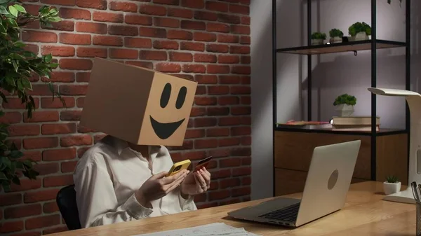 Business life and office daily routine creative advertisement concept. Portrait of female in cardboard box with emoji on head. Worker at the desk holding smartphone and credit card typing number.