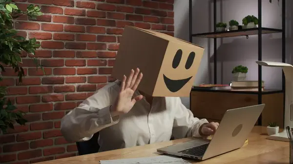 Business life and office daily routine creative advertisement concept. Portrait of female in cardboard box with emoji on head. Worker sitting at the desk talking by video call on laptop, happy face.