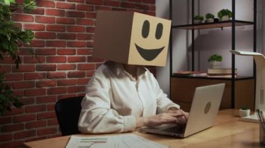 İş hayatı ve ofis günlük yaratıcı reklam konsepti. Kafasında emoji olan karton kutuda bir kadın portresi. Resepsiyonda çalışan biri dizüstü bilgisayarda çalışıyor, el ele tutuşup kameraya bakıyor..