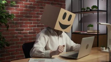 İş hayatı ve ofis günlük yaratıcı reklam konsepti. Kafasında emoji olan karton kutuda bir kadın portresi. Masada oturan bir işçi dizüstü bilgisayarda çalışıyor, mutlu bir ifade var..