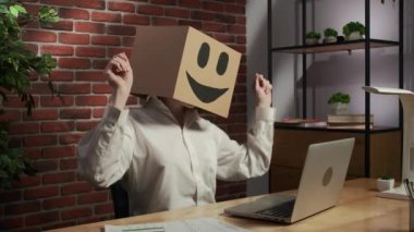 İş hayatı ve ofis günlük yaratıcı reklam konsepti. Kafasında emoji olan karton kutuda bir kadın portresi. İşçi masada oturuyor, dizüstü bilgisayarda çalışıyor, bitiriyor ve dans etmeye başlıyor..