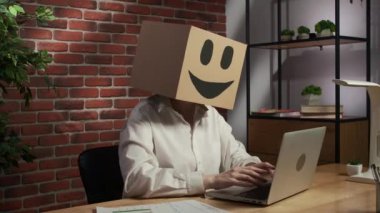 İş hayatı ve ofis günlük yaratıcı reklam konsepti. Kafasında emoji olan karton kutuda bir kadın portresi. İşçi masada oturuyor dizüstü bilgisayarla çalışıyor, sandalyeye yaslanıyor..