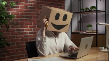 İş hayatı ve ofis günlük yaratıcı reklam konsepti. Kafasında emoji olan karton kutuda bir kadın portresi. İşçi masada oturuyor dizüstü bilgisayarla çalışıyor, akıllı telefonla konuşuyor..