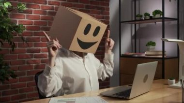 İş hayatı ve ofis günlük yaratıcı reklam konsepti. Kafasında emoji olan karton kutuda bir kadın portresi. İşçi masada oturmuş dizüstü bilgisayara bakıyor. Mutlu dans havası.