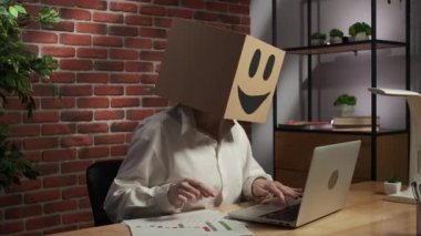 İş hayatı ve ofis günlük yaratıcı reklam konsepti. Kafasında emoji olan karton kutuda bir kadın portresi. Masada oturan işçiler kağıt belgelerle meşgul dizüstü bilgisayarlar üzerinde çalışıyorlar..
