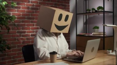 İş hayatı ve ofis günlük yaratıcı reklam konsepti. Kafasında emoji olan karton kutuda bir kadın portresi. Masada oturan bir işçi dizüstü bilgisayarda çalışıyor, elinde bir fincan kahve tutuyor.,