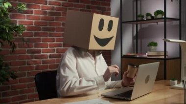 İş hayatı ve ofis günlük yaratıcı reklam konsepti. Kafasında emoji olan karton kutuda bir kadın portresi. İşçi masada oturup laptopla çalışıyor ve bir fincan kahve içiyor.,