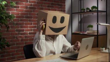 İş hayatı ve ofis günlük yaratıcı reklam konsepti. Kafasında emoji olan karton kutuda bir kadın portresi. Çalışma masasında çalışan biri akıllı telefondan konuşuyor, kafası karışmış ifadeyi kapatıyor..