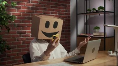 İş hayatı ve ofis günlük yaratıcı reklam konsepti. Kafasında emoji olan karton kutuda bir kadın portresi. Masada çalışan bir işçi akıllı telefondan konuşuyor duygusal olarak, üzgün suratlı bir kutuya dönüşüyor.
