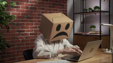 İş hayatı ve ofis günlük yaratıcı reklam konsepti. Kafasında emoji olan karton kutuda bir kadın portresi. Masada oturan işçiler dizüstü bilgisayar üzerinde çalışıyorlar. Üzgün ifadeler..