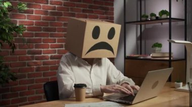İş hayatı ve ofis günlük yaratıcı reklam konsepti. Kafasında emoji olan karton kutuda bir kadın portresi. Çalışma masasında oturan üzgün suratlı, elinde kahve fincanı tutan bir işçi.