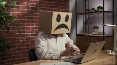 İş hayatı ve ofis günlük yaratıcı reklam konsepti. Kafasında emoji olan karton kutuda bir kadın portresi. Masada oturan işçiler dizüstü bilgisayarda çalışıyor, üzgün yüzler soğuk iç geçiriyor.