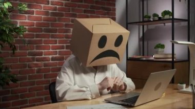 İş hayatı ve ofis günlük yaratıcı reklam konsepti. Kafasında emoji olan karton kutuda bir kadın portresi. İşçi masada oturmuş laptoptaki video görüşmesiyle konuşuyor. Üzücü bir duygu..