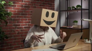 İş hayatı ve ofis günlük yaratıcı reklam konsepti. Kafasında emoji olan karton kutuda bir kadın portresi. İşçi masada oturmuş bilgisayardan video görüşmesi yapıyor, gülen surat..