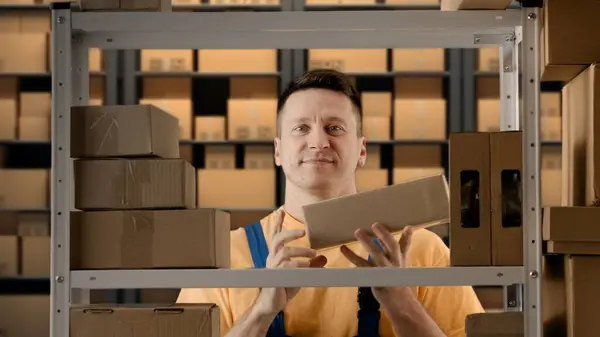 Business warehouse and logistics creative advertisement concept. Portrait of male working in storage. Man storekeeper standing near rack with boxes, holding package in hand shows thumbs up at camera.